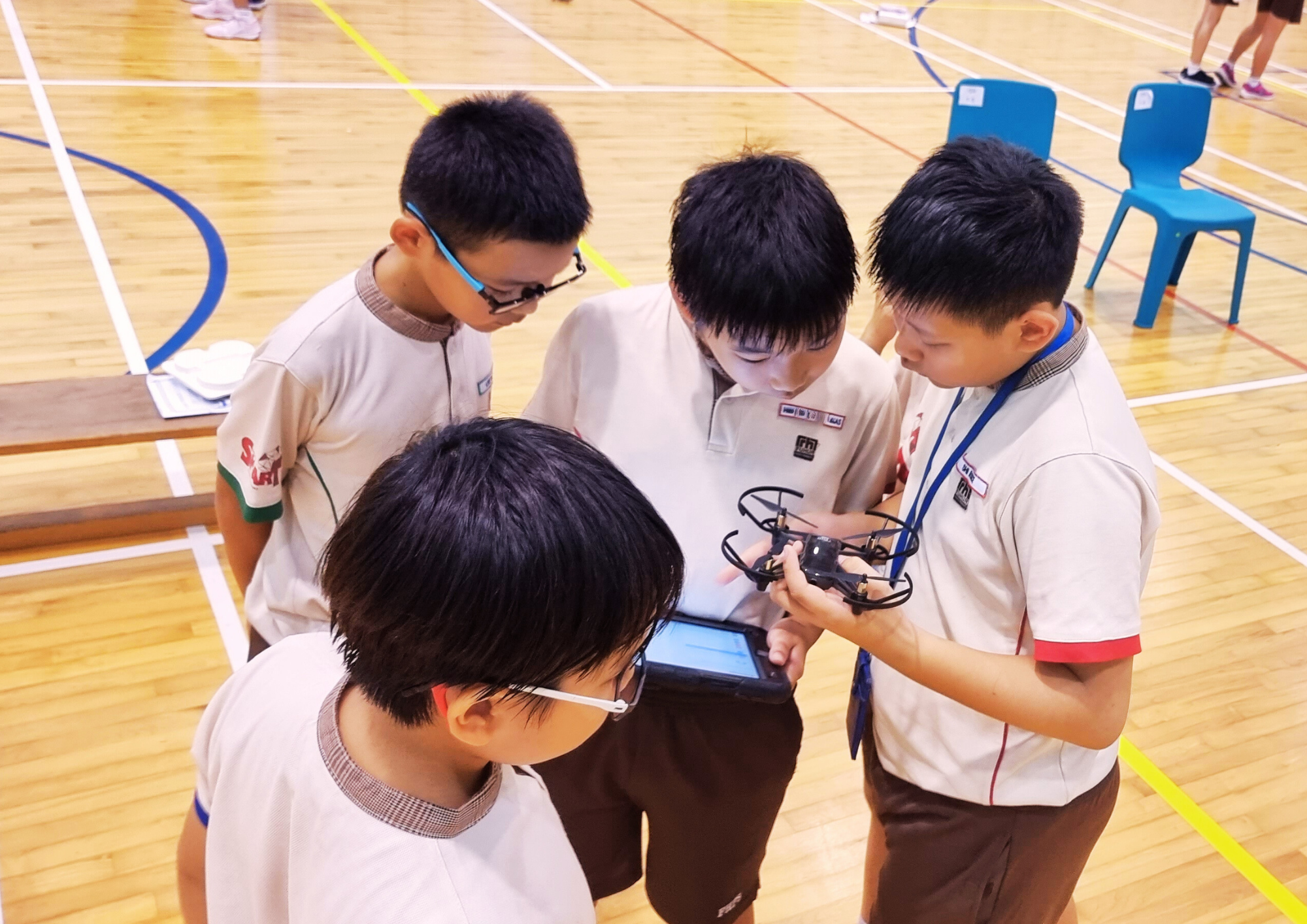 Four students in a gym huddle over a tablet while one holds a small quadcopter drone.